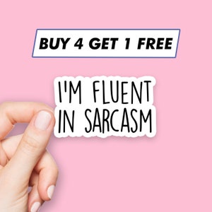 Könnte beinhalten: Weißer Aufkleber mit schwarzem Text, der "Ich bin flüssig im Sarkasmus" lautet. Eine Hand hält den Aufkleber. Der Hintergrund ist rosa. Der Text "KAUFE 4, BEKOMME 1 GRATIS" befindet sich in einem blauen Rechteck oben im Bild.