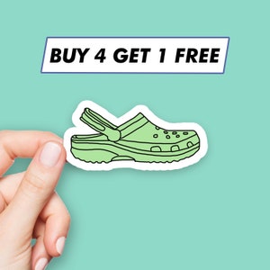 Puede incluir: Una pegatina verde claro de un zapato Croc con borde blanco. La pegatina está sujeta por una mano sobre un fondo turquesa. Encima del zapato hay un rectángulo blanco con el texto "BUY 4 GET 1 FREE" en negro.