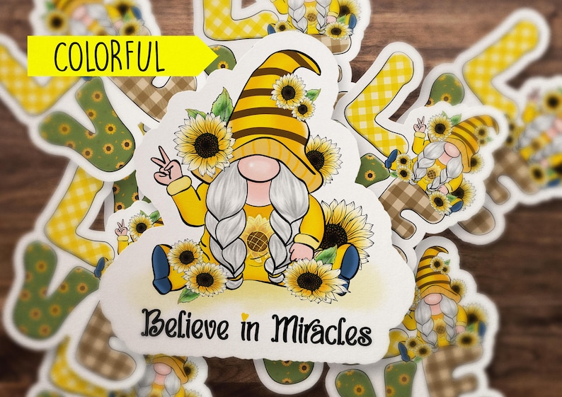 May include: A white sticker with a yellow and brown gnome wearing a sunflower crown and holding a peace sign. The gnome is surrounded by sunflowers and the sticker says "Believe in Miracles".
