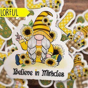 May include: A white sticker with a yellow and brown gnome wearing a sunflower crown and holding a peace sign. The gnome is sitting on a bed of sunflowers. The sticker says "Believe in Miracles".