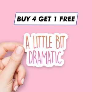 Puede incluir: Pegatina blanca con el texto "A little bit dramatic" en letras con degradado rosa y naranja. La pegatina está siendo sostenida por una mano sobre un fondo rosa.