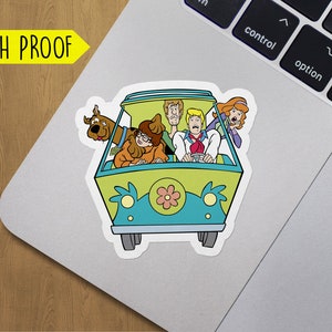 May include: A sticker featuring the Mystery Machine from Scooby-Doo. The van is green and blue with a yellow flower on the front. The characters Scooby-Doo, Shaggy, Fred, Daphne, and Velma are inside the van.