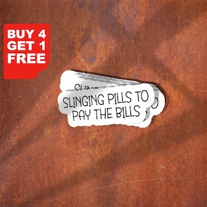 Op de afbeelding: Een stapel witte stickers met de tekst "SLINGING PILLS TO PAY THE BILLS" in zwart, met een rode hoek met de tekst "BUY 4 GET 1 FREE" in wit. De stickers liggen op een roestbruine achtergrond.