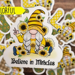 May include: A white sticker with a yellow and brown gnome wearing a sunflower crown and holding a peace sign. The gnome is sitting on a bed of sunflowers. The sticker says "Believe in Miracles".