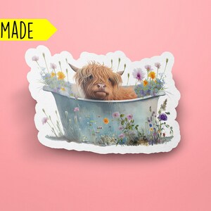 K&ouml;nnte beinhalten: Ein handgemachter Sticker mit einer Highland-Kuh, die in einer mit Wildblumen gef&uuml;llten Wanne ein Bad nimmt. Die Kuh ist braun und wei&szlig; mit einem flauschigen Fell. Die Wanne ist blau und wei&szlig; mit einem rustikalen Look. Die Blumen sind in verschiedenen Farben, darunter rosa, lila, gelb und wei&szlig;.