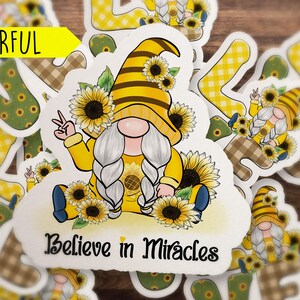May include: A colorful sticker featuring a gnome with a yellow and black striped hat, holding a peace sign. The gnome is surrounded by sunflowers and the text "Believe in Miracles".