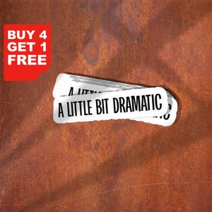Puede incluir: Pegatina blanca con el texto "A LITTLE BIT DRAMATIC" en negro, apilada sobre una superficie marrón oxidada texturizada. Un gráfico de esquina roja dice "BUY 4 GET 1 FREE" en blanco.
