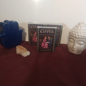 May include: Three decks of Fin de Siècle Kipper Fortune Telling Cards, with a green and gold design, featuring a couple in a romantic setting. The cards are on a red surface with a blue hamsa hand, a white crystal, and a white ceramic Buddha statue.