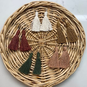 May include: A collection of handmade tassel earrings in various colors, including white, brown, green, and pink. The earrings are displayed on a woven wicker tray. Each pair features a looped top and a long, fringed tassel.