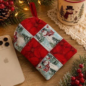 May include: A quilted fabric pot holder featuring a patchwork design of red poinsettia flowers and a winter scene with snowmen, pine trees, and red birds. The square-shaped kitchen accessory includes a small fabric loop for hanging on the top edge.