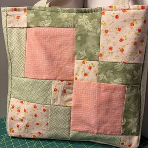 May include: A patchwork tote bag with a cream-colored handle. The bag features a mix of floral, geometric, and solid fabric squares in shades of pink, green, and cream. The bag is made of quilted fabric.