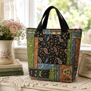 May include: A quilted tote bag featuring a patchwork design with various floral and paisley patterns in shades of black, green, orange, and blue. The bag has two black fabric handles and a zippered top closure, constructed with a stitched grid pattern.