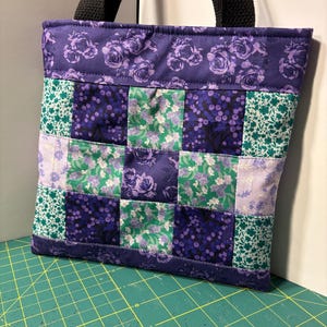 May include: A quilted tote bag with a patchwork design. The bag features a combination of purple floral and green floral fabrics. The top and bottom edges are trimmed with a solid purple fabric. The bag has black handles.