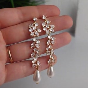 May include: Gold-coloured drop earrings with a floral design. Each earring features clear crystals and a pear-shaped faux pearl. The earrings have a cascading vine-like pattern, suitable for special events.