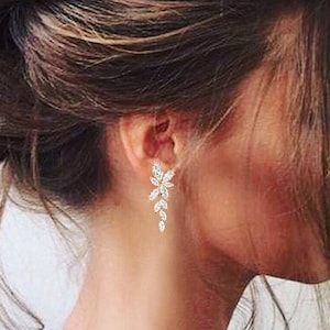 Puede incluir: Una mujer con el pelo largo y castaño recogido en un moño, lleva un par de pendientes colgantes con diseño de hojas y piedras transparentes.