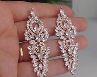 May include: A pair of long, rose gold earrings with a teardrop design. The earrings are adorned with clear, sparkling crystals.