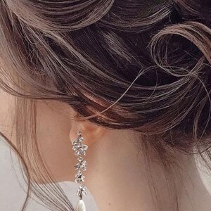 May include: A pair of elegant drop earrings with a floral design, clear crystals, and a pearl drop. The earrings are set against dark brown hair. A stylish accessory for formal events, adding a touch of glamour to any outfit.