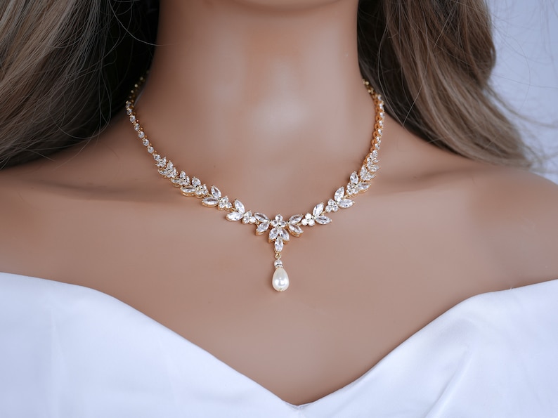 May include: A gold-coloured necklace adorned with clear, sparkling crystals and a pear-shaped pearl pendant. The necklace showcases a leaf-like design with a delicate arrangement of crystals. The necklace is worn on a person wearing a white top.