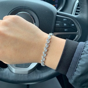 May include: A silver bracelet featuring a leaf pattern, set with clear, sparkling stones. The bracelet is worn on a wrist, with a black sleeve visible. The background includes a car's steering wheel and controls.