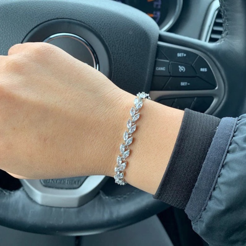 May include: A silver bracelet featuring a leaf pattern, set with clear, sparkling stones. The bracelet is worn on a wrist, with a black sleeve visible. The background includes a car's steering wheel and controls.
