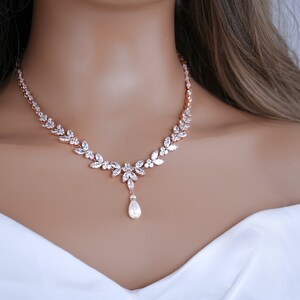 May include: Rose gold necklace with a floral design, set with clear crystals. The necklace has a central pendant with a pear-shaped pearl drop. The jewellery is displayed on a mannequin.