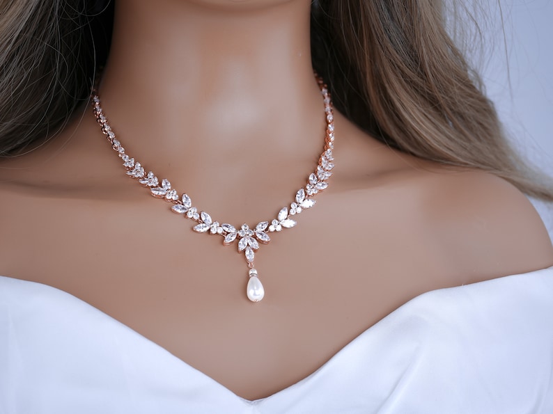 May include: Rose gold necklace with a floral design, set with clear crystals. The necklace has a central pendant with a pear-shaped pearl drop. The jewellery is displayed on a mannequin.