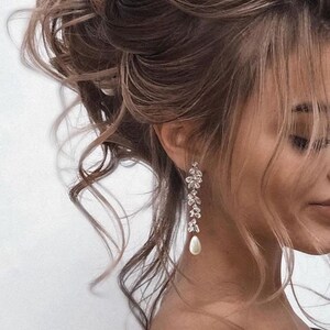 May include: Silver-toned drop earrings featuring a floral design and a pear-shaped pearl. The earrings are shown in close-up, with a person's hair styled in an updo. The earrings dangle elegantly.