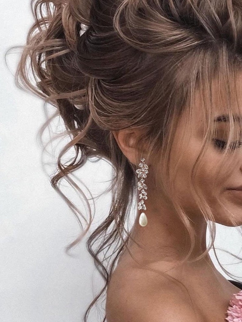 May include: Silver-toned drop earrings featuring a floral design and a pear-shaped pearl. The earrings are shown in close-up, with a person's hair styled in an updo. The earrings dangle elegantly.