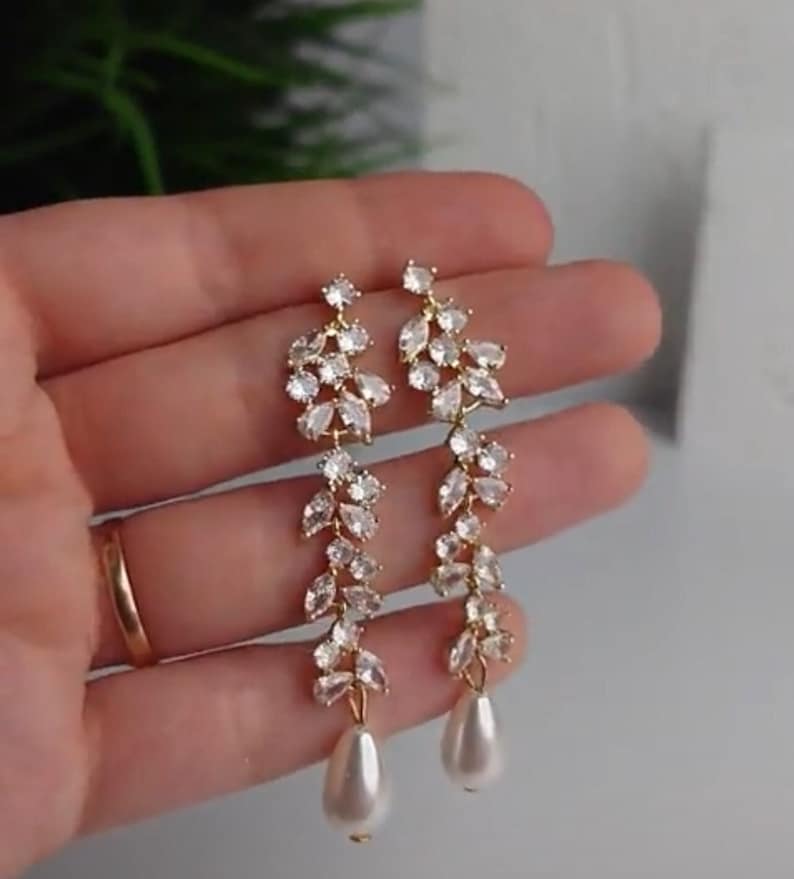May include: Gold-coloured drop earrings with a floral design. Each earring features clear crystals and a pear-shaped faux pearl. The earrings have a cascading vine-like pattern, suitable for special events.