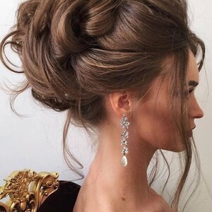 May include: A woman's hair is styled in a voluminous updo of dark brown curls. She wears long, dangling earrings with floral designs and a pear-shaped pearl drop. The earrings are silver-toned, adding a touch of elegance.