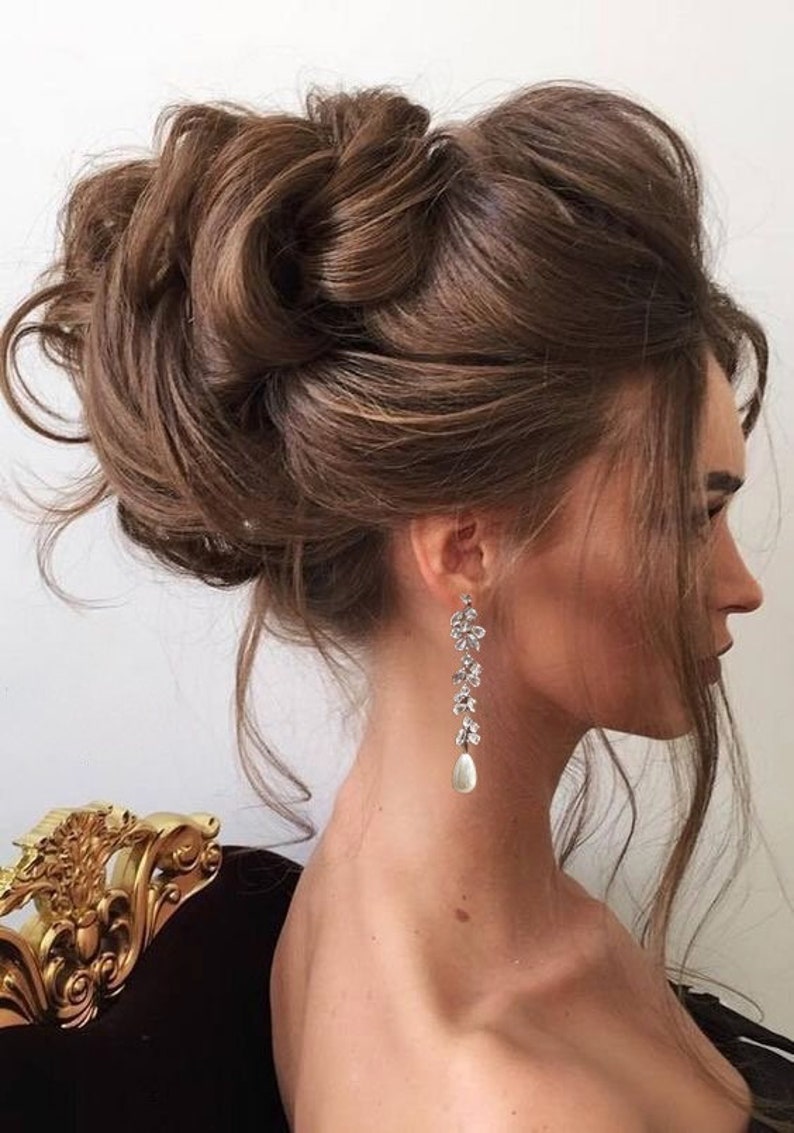 May include: A woman's hair is styled in a voluminous updo of dark brown curls. She wears long, dangling earrings with floral designs and a pear-shaped pearl drop. The earrings are silver-toned, adding a touch of elegance.