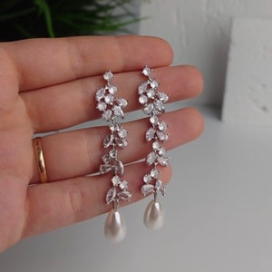 May include: Silver-coloured earrings featuring a leaf design, embellished with clear crystals and a pear-shaped pearl drop. The earrings are long and dangle. The crystals sparkle, adding a touch of glamour. The earrings are a stylish accessory.