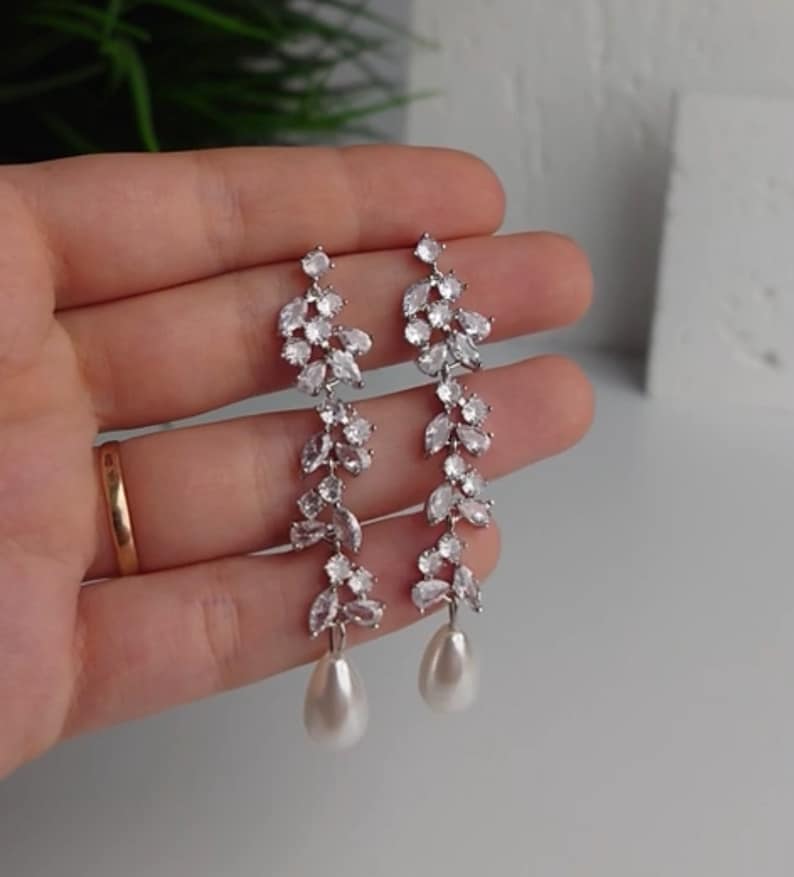 May include: Silver-coloured earrings featuring a leaf design, embellished with clear crystals and a pear-shaped pearl drop. The earrings are long and dangle. The crystals sparkle, adding a touch of glamour. The earrings are a stylish accessory.