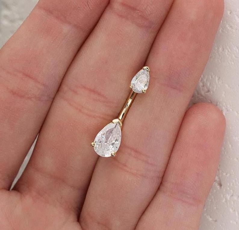 Puede incluir: Un anillo de ombligo de oro con dos piedras transparentes en forma de l&aacute;grima.