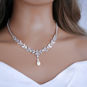 May include: A silver necklace with a floral design. The necklace features clear, sparkling crystals and a pear-shaped pearl pendant. The necklace is worn on a person wearing a white top.