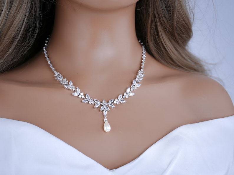 May include: A silver necklace with a floral design. The necklace features clear, sparkling crystals and a pear-shaped pearl pendant. The necklace is worn on a person wearing a white top.