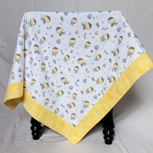 May include: A white baby blanket with a repeating pattern of cartoon bees and beehives. The blanket has a yellow border with a floral design. The blanket is approximately 40 inches square.