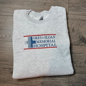 May include: Grey's Anatomy inspired sweatshirt with the embroidered text "GREY+SLOAN MEMORIAL HOSPITAL" in blue and red on a grey background.