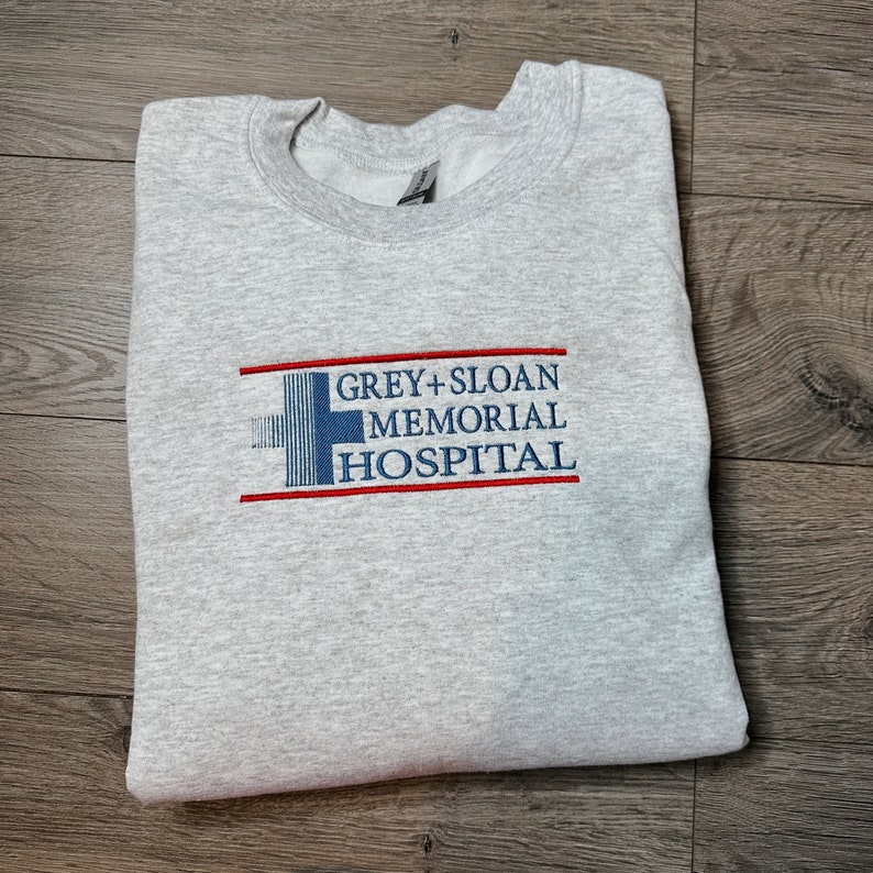 May include: Grey's Anatomy inspired sweatshirt with the embroidered text "GREY+SLOAN MEMORIAL HOSPITAL" in blue and red on a grey background.