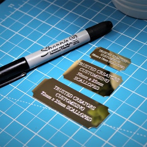 May include: Three gold-coloured metal plaques with scalloped edges and the text "Trusted Creators Customising" followed by the dimensions and "Scalloped". The plaques are on a blue cutting mat.