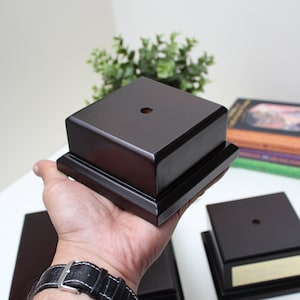 May include: A dark brown wooden trophy base with a square top and a small hole in the centre. The base has a raised edge around the top and bottom.