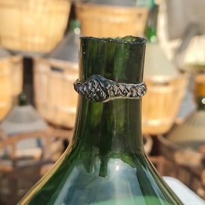 Può includere: Una bottiglia di vetro verde con un anello decorativo in metallo scuro attorno al collo. La bottiglia è a fuoco, con altre bottiglie simili e cesti di vimini sullo sfondo. La bottiglia ha una base larga e un collo stretto.