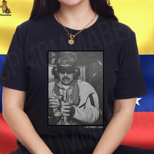 Peut inclure: T-shirt noir avec une image en noir et blanc d'un homme portant un casque et des lunettes de soleil, tenant des objets. Le texte "3 DE ENERO, 2026" est en dessous de l'image. Un collier doré est porté.