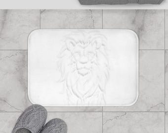 May include: A white rectangular bath mat with an embossed lion's head design. The mat is on a tiled floor, with a gray towel and a pair of gray knitted slippers. The mat is a bathroom accessory, suitable for a modern bathroom.