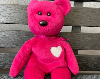 May include: A pink beanie baby with a white heart on its chest. The beanie baby is sitting on a gray textured surface.