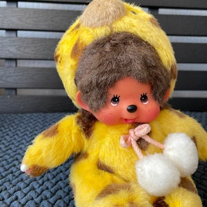 May include: A plush toy monkey wearing a yellow and brown giraffe costume with white pom-poms on its feet.