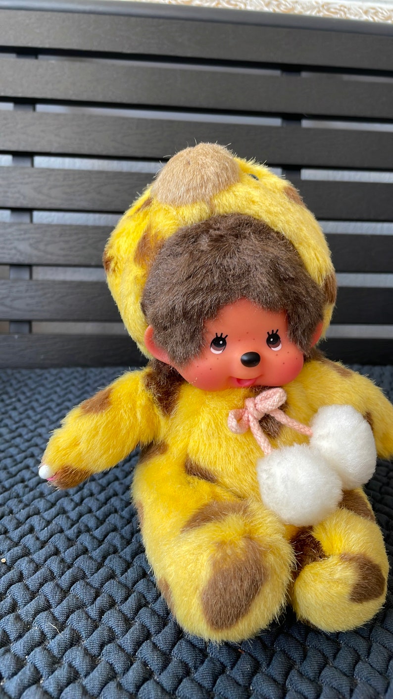 May include: A plush toy monkey wearing a yellow and brown giraffe costume with white pom-poms on its feet.