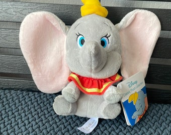 May include: A gray plush toy elephant with a yellow hat and red and yellow striped collar. The elephant is sitting on a gray textured surface. The toy is labeled "Disney Classic Plush Collection".