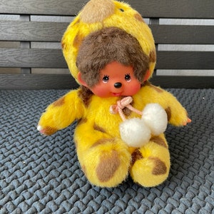 May include: A yellow and brown plush toy monkey wearing a giraffe costume with white pom-poms. The monkey has brown fur, a pink nose, and black eyes.
