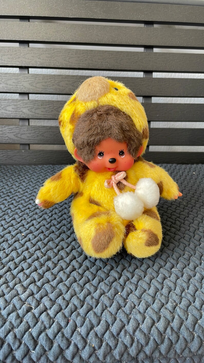 May include: A yellow and brown plush toy monkey wearing a giraffe costume with white pom-poms. The monkey has brown fur, a pink nose, and black eyes.